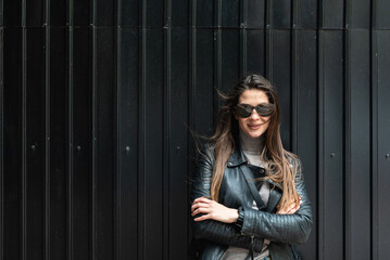 Fototapeta premium Portrait of young beautiful cool looking woman in leather jacker and sunglasses standing against black wall. Cute female positive attitude and crossed hands posing outdoor while wing wave her hair.
