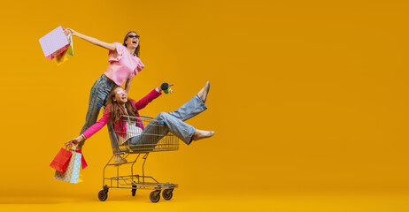 Two young excited girls go shopping with shopping cart isolated on bright yellow background. Concept of sales, black friday, discount, emotions - obrazy, fototapety, plakaty
