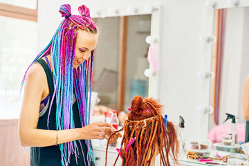Hippie style hair. Woman hairdresser with colored afro braids weaves to girl ginger dreadlocks. Stylish therapy professional care concept. Weaving process with kanekalon. Beauty salon services.