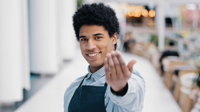 Friendly african american young guy male waiter salesman restaurant cafe kitchen bar worker in apron looking at camera showing gesture her you come here approach welcome inviting customers promotions