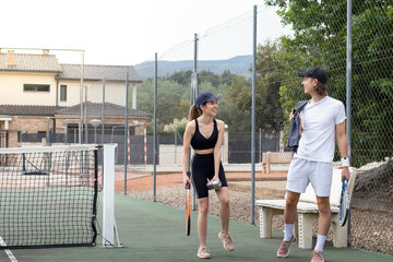 Man and woman looking to each other arriving to a tennis court with their racquets while laughing and talking