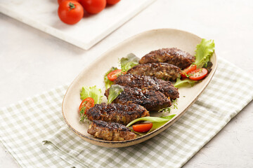 Several homemade roasted minced meat sausages köfte on an oval beige colored plate with green leaves and cherry tomatoes on green checkered kitchen towel