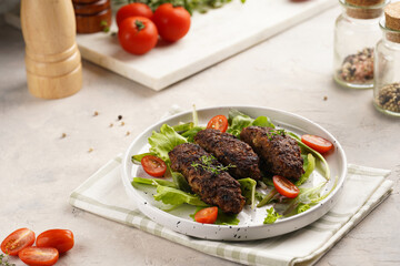 Several homemade roasted minced meat sausages köfte on a round white plate with green leaves and cherry tomatoes on beige colored linen napkin