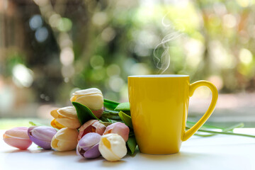 Yellow steaming cup of hot coffee on white wooden near window with tulip flowers and green nature blurred background.