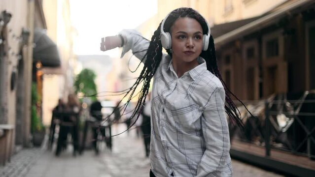 Young beautiful woman with dreadlocks listening music and dancing in the city centre. Slow motion of happy young woman in headphones dancing outdoors in city street.