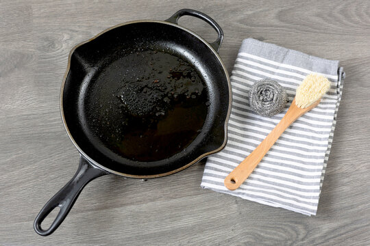 Dirty Cast Iron Skillet Being Prepared For Cleaning With Bamboo Brush, Stainless Steel Scrubber And Dish Towel On A Gray Wood Counter.