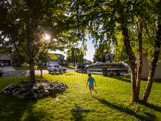 A boy runs through a sprinkler in a suburban yard with the sun shining through the trees on a summer day - obrazy, fototapety, plakaty