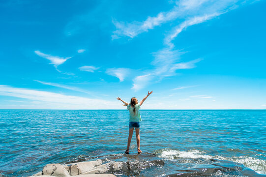 A girl stands on a rocky outcrop in front of a Lake Superior and a bright blue sky with her hands in the air 