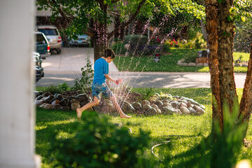 A young boy runs through a sprinkler in a suburban neighborhood on a summer day - obrazy, fototapety, plakaty