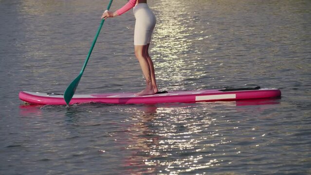 Young woman with slender legs floating on paddle board during amazing sunset. Female ahlete in sprit clothes having evenin gworkout on fesh air. Close up.