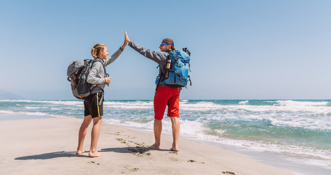 Father and teenager son with backpacks making High FIVE on sandy seaside beach during Lycian Way trekking walk. Famous Likya Yolu Turkish route. Active happy family people vacation concept image.