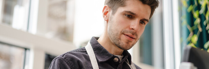 Obraz premium Serious young man in apron standing at the cash register