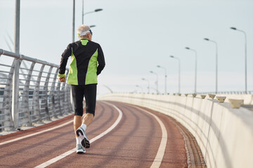 Horizontal back view shot of senior man wearing black and green tracksuit running along track