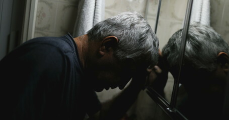 Desperate senior leaning head on bathroom mirror suffering from illness