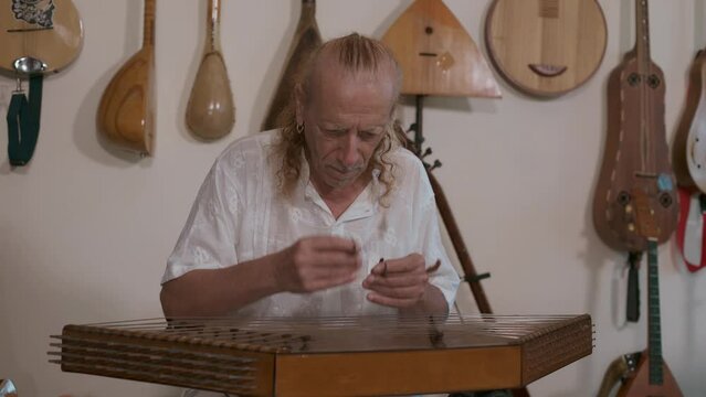 Elderly caucasian man playing traditional musical instrument of Middle East santur in his home studio. Behind him on the wall is a collection of oriental stringed musical instruments.