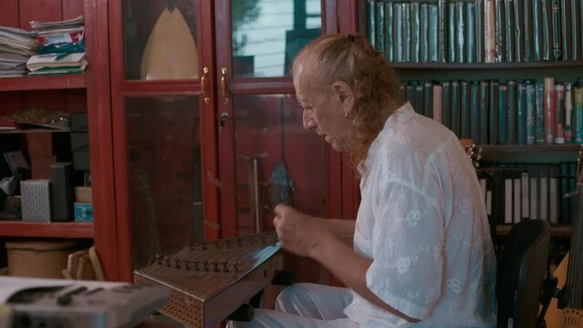 Elderly caucasian man playing traditional musical instrument of Middle East santur in his home studio. Behind him on the wall is a collection of oriental stringed musical instruments.