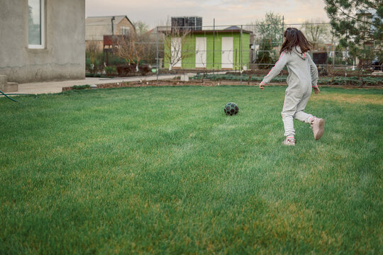 Cute little girl playing football with soccer ball on green lawn in backyard of house. Child kicking soccer ball on field. High quality photo