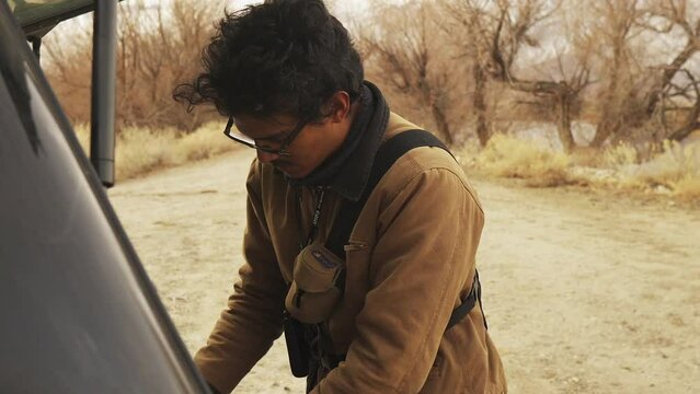 Medium shot of a man with unkempt black hair, thick framed glasses and a mustache packing and preparing equipment in the back of a pickup truck with a covered bed, in the  high desert talking