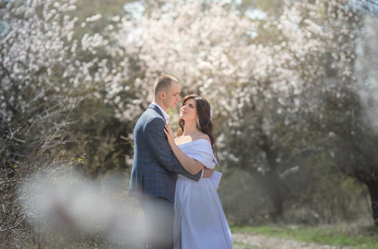 a guy in a suit and a girl in a white dress are standing in a garden with flowering trees and hugging.