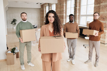 Fototapeta na wymiar Portrait of content ambitious young lady in casual suit holding moving box against male colleagues in modern office