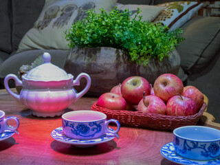 Table set with coffe cups, sugar bowl, a basket of red apples and a potted plant.