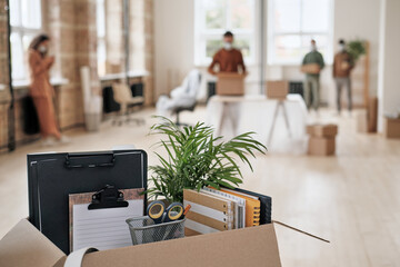 Focus on open cardboard box with office supplies, clipboards and plant against people moving into...