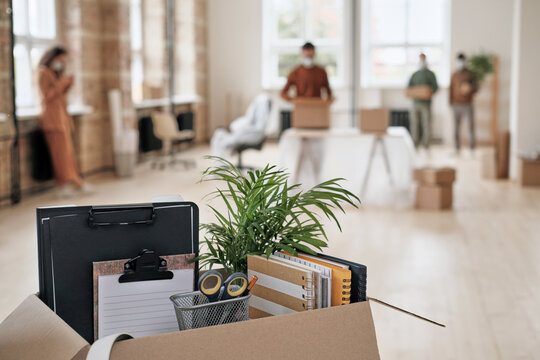 Focus on open cardboard box with office supplies, clipboards and plant against people moving into new office in background