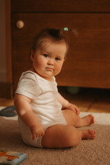 A 7-month girl is sitting near a dresser in a bodysuit at home.