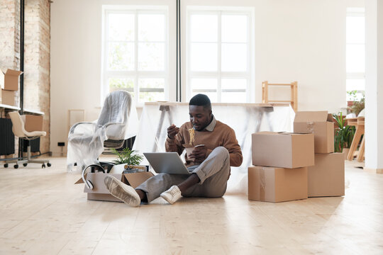 Too Busy Young Black Manager Sitting With Laptop On Floor And Eating Noodles At Workplace To Finish Project On Deadline In Office With Moving Boxes