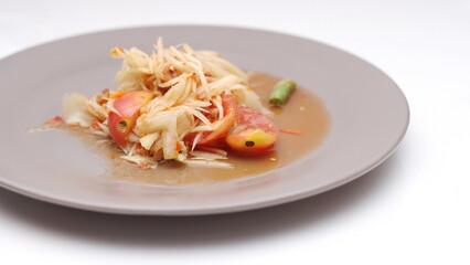 Thai papaya salad on a plate, papaya salad with poop, lime, pepper, red tomatoes in a gray plate on a white table.