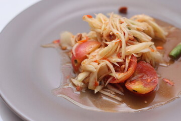 Thai papaya salad on a plate, papaya salad with poop, lime, pepper, red tomatoes in a gray plate on a white table.