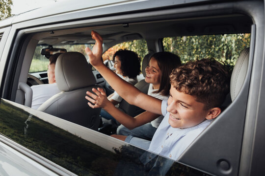 Teenage Boy and Girl Greeting to Someone Through Window with Waving Hands While Sitting Inside Minivan Car, Happy Four Members Family on a Weekend Road Trip