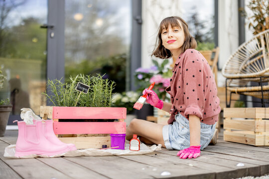 Young woman painting wooden box in pink color, doing some renovating housework on the terrace outdoors. DIY concept