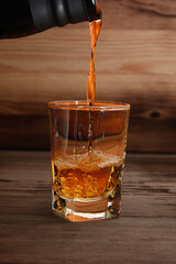 Alcoholic strong drink whiskey is poured from a bottle into a glass. Close-up on a wooden background