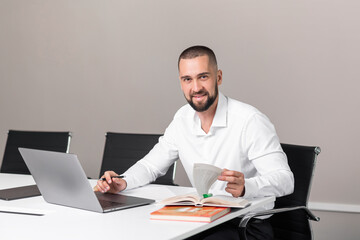 Handsome strong young man with beard in white shirt with working in office with books and laptop