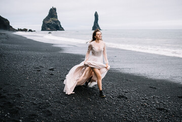 Beautiful young girl in wedding dress and black boots run on Black Sand beach in Vic, Iceland with high rock islands in background
