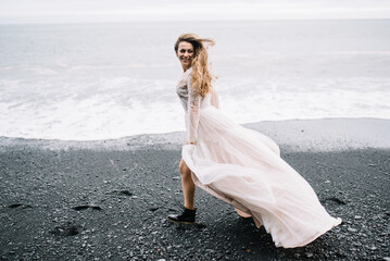 Fototapeta na wymiar Young girl in wedding dress and black boots run on Black Sand beach in Vic, Iceland 