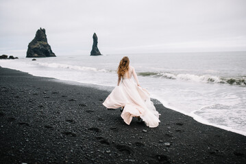 Girl in old old-styled wedding dress are running on the Black Sand beach in Vic, Iceland with rock...