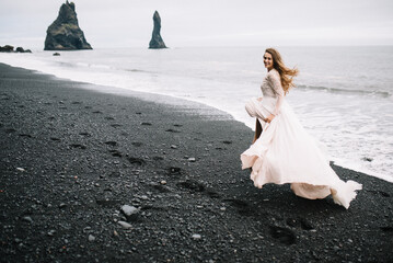 Fototapeta na wymiar Bride in old classy wedding dress are running out on the Black Sand beach in Vic, Iceland with rock islands on background and waves 