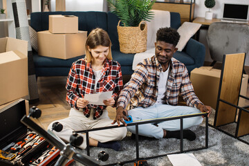 Multicultural young couple measuring shelf length while reading instructions indoors. Smiling...