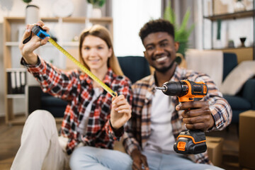 Fototapeta na wymiar Blur background of multi ethnic couple in casual clothes showing measure tape and electric drill on camera. Cheerful family using tools for repairing and assembling furniture at new house.