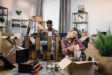 Positive woman unpacking boxes and talking on mobile phone while man assembling furniture behind. Young multiracial couple enjoying moving process to new house.