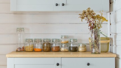A row of glass jars of groceries, a bouquet of dried flowers and a mesh square basket are on the table. Scandinavian design idea, food storage, zero waste