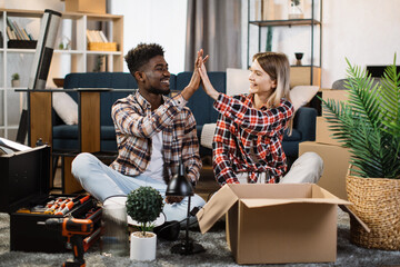 Handsome african american man and beautiful caucasian woman making high five gesture while sitting on floor at living room with boxes around. Concept of family and relocation.