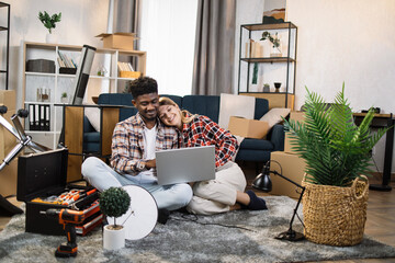 African american man and caucasian woman sitting in embrace on floor and using modern laptop at home. Smiling young couple searching furniture online for new home.
