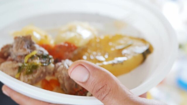 Man's hands holding a plastic bowl and a metal spoon pour hot boiled Armenian khashlama from the cauldron with delicious juicy pieces of beef meat and potatoes