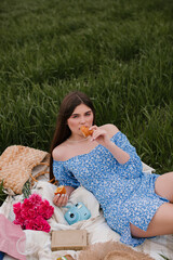 A beautiful girl with long hair in a blue dress eats a croissant at a picnic in nature.