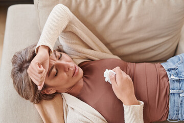 Horizontal from above shot of modern woman suffering headache and runny nose relaxing on sofa at home