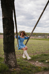 A girl with long dark hair, in a blue dress, white boots and a straw hat, rides on a swing in nature.