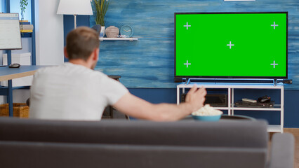 Sports fan watching game on green screen tv mockup encouraging favourite team while relaxing at...